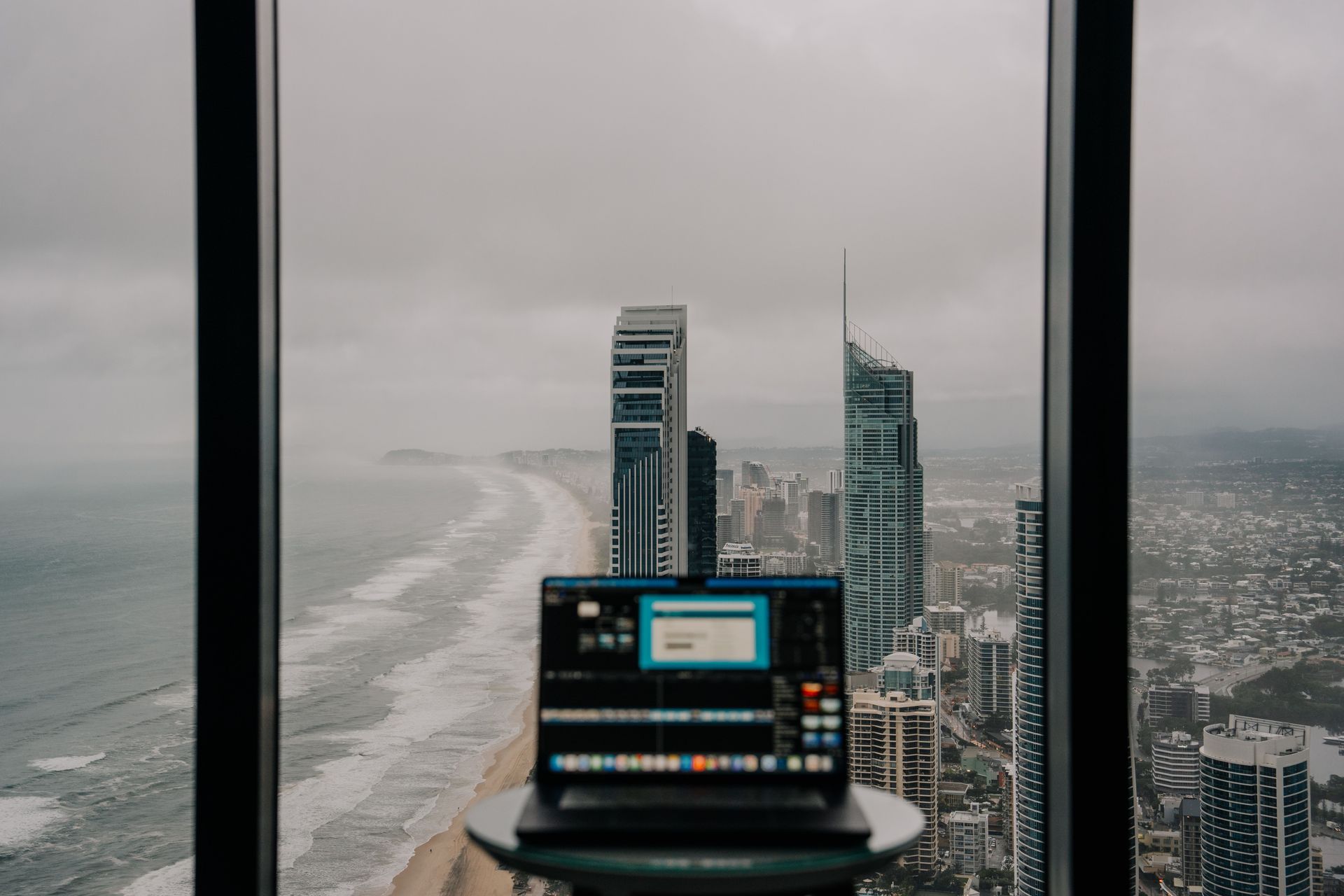 Laptop on a table overlooking a rainy city skyline through large windows.