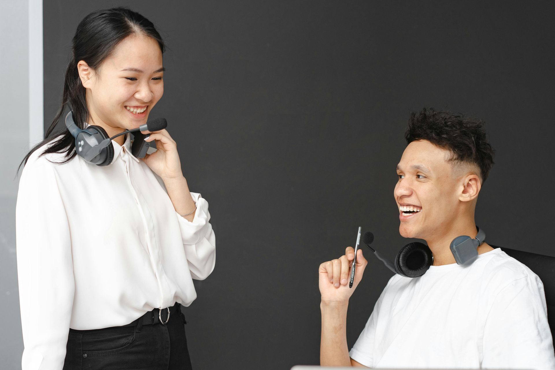 Two coworkers smiling with headsets, one holding a pen, against a dark background