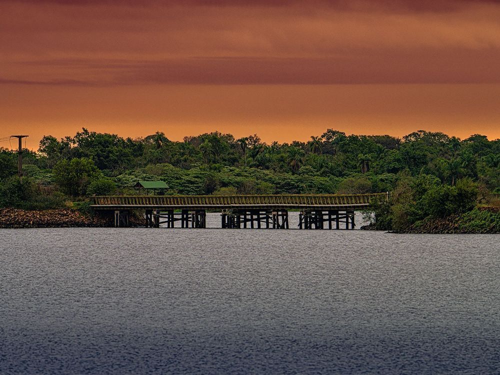 Puente de madera sobre aguas tranquilas al atardecer, con frondosos árboles verdes bajo un cielo anaranjado.