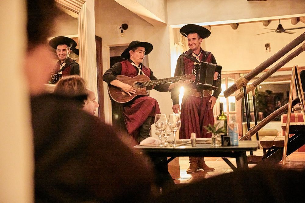 Tres músicos vestidos con trajes rojos actúan en un escenario con iluminación cálida; uno de ellos toca la guitarra junto a una mesa iluminada por una linterna.