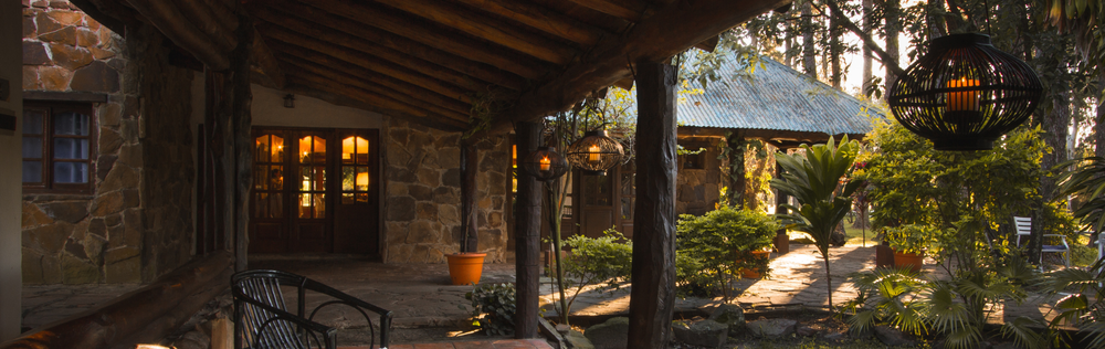 Porche rústico de cabaña con ventanas cálidamente iluminadas, farol colgante y un sendero de jardín bañado por el sol al atardecer.
