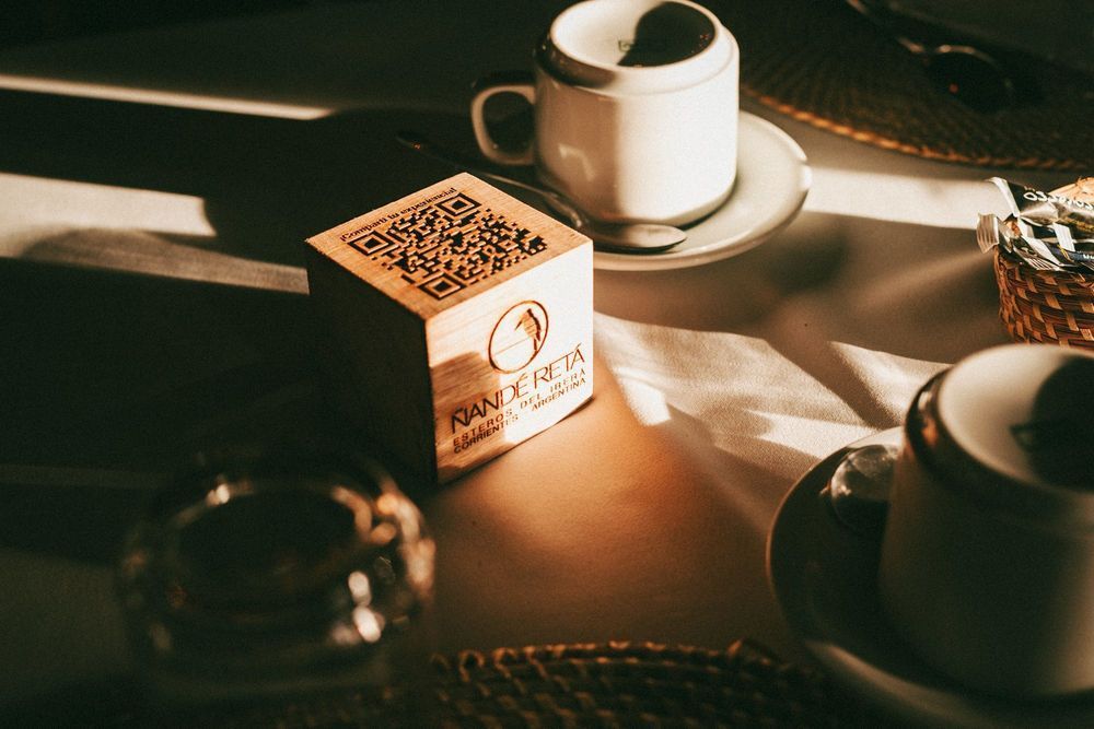 Pequeño cubo de madera con código QR junto a tazas de café en una mesa iluminada por el sol.