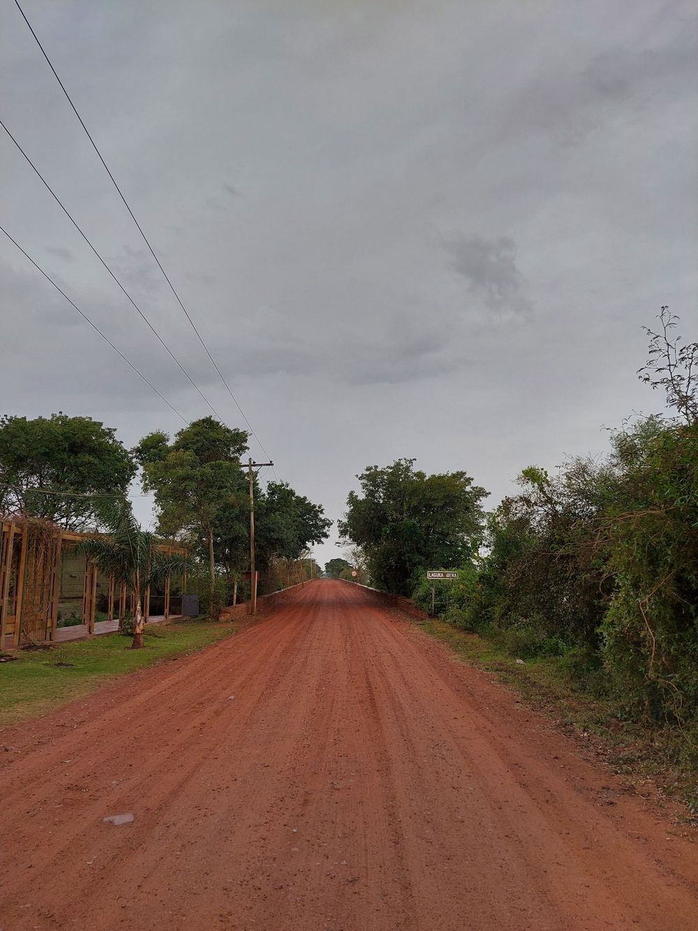 Un camino de tierra roja se extiende hasta el horizonte, bordeado de árboles y líneas eléctricas bajo un cielo nublado.
