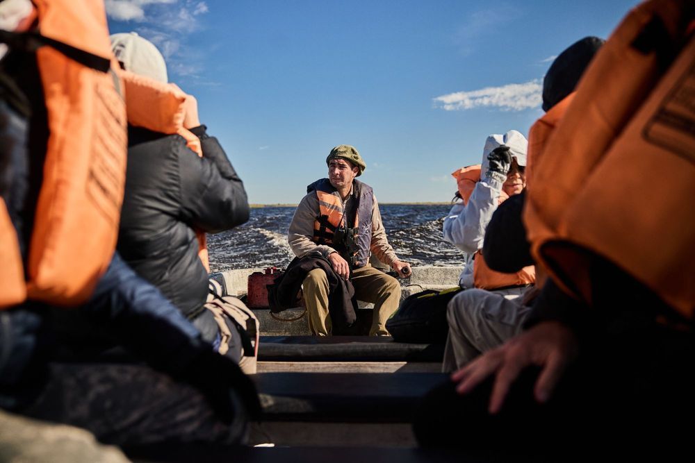 Un grupo de personas con chalecos salvavidas naranjas navega en una pequeña embarcación en aguas abiertas bajo un cielo azul despejado.