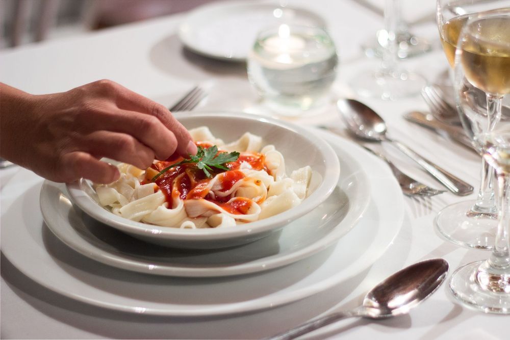 Una mano se extiende para alcanzar la pasta sobre un mantel blanco, con copas de vino y cubiertos puestos para la cena.