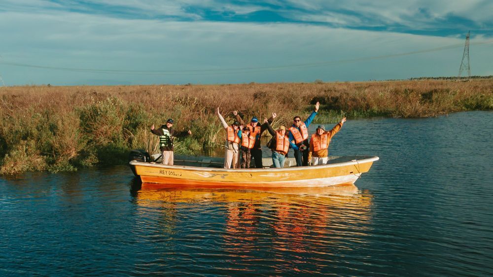 En una barca naranja, la gente alza los brazos en un río tranquilo junto a los juncos al atardecer.