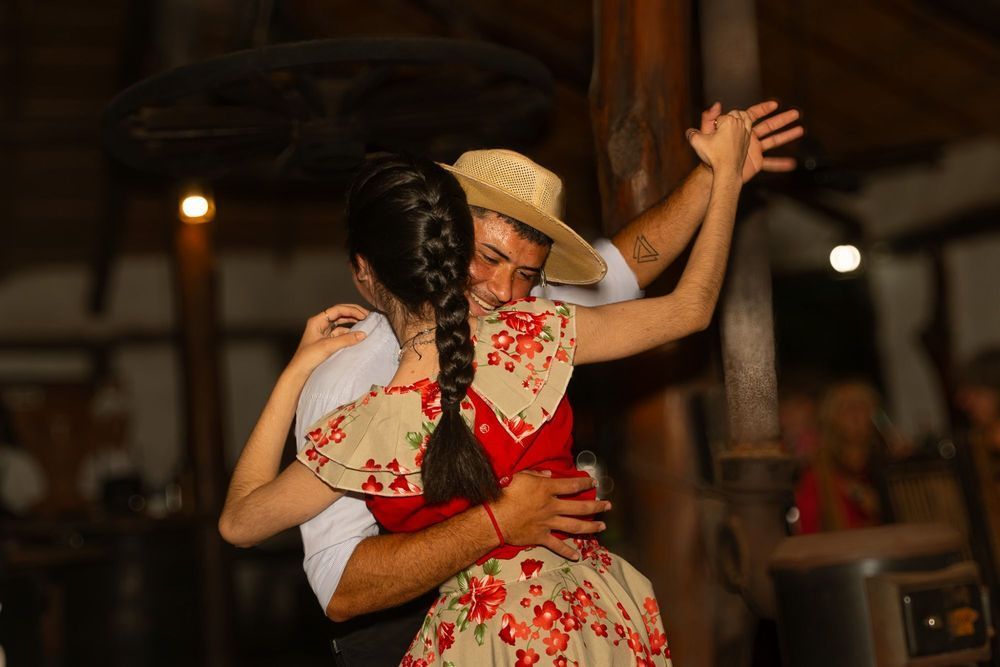 Una pareja baila muy junta en un local rústico con poca luz; la mujer lleva un vestido de flores y el hombre un sombrero de paja.