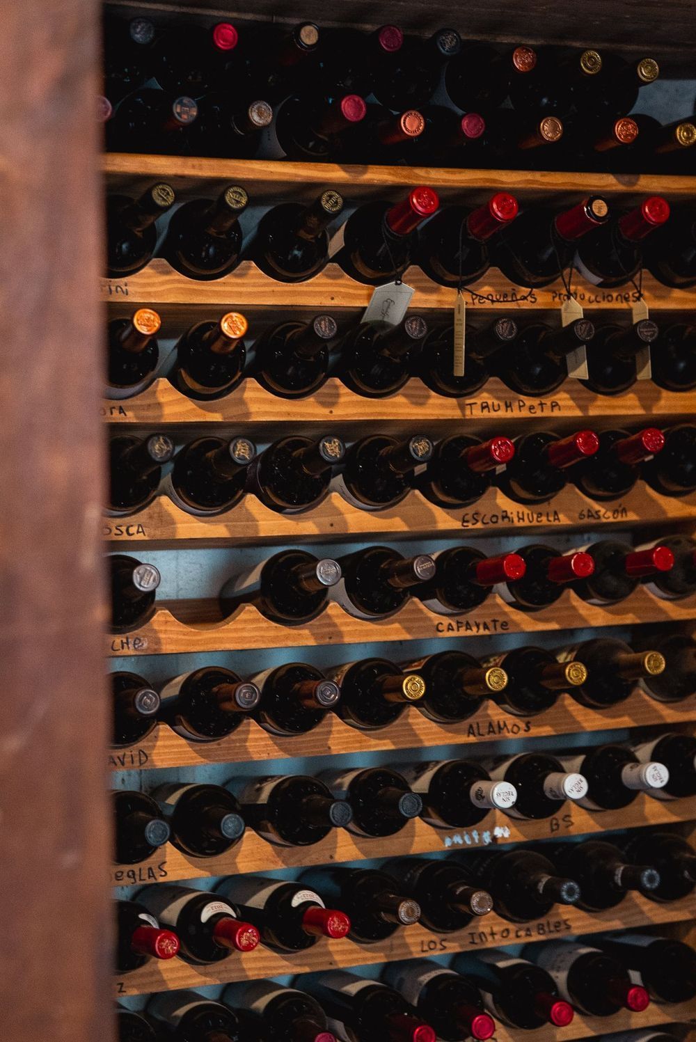 Botellas de vino almacenadas en un estante de madera, con filas de cuellos de botella y etiquetas de colores visibles.