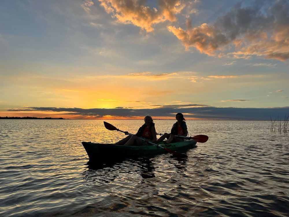 Dos personas reman en kayak sobre aguas tranquilas al atardecer, bajo un cielo nublado y colorido.