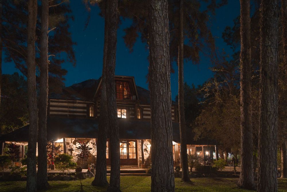Una casa cálidamente iluminada por la noche entre árboles altos, con grandes ventanales que brillan contra un cielo oscuro.