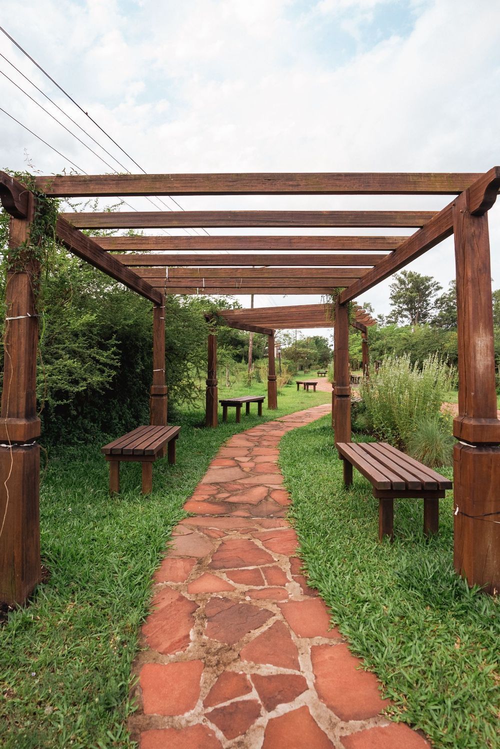 Sendero rústico de jardín con camino de piedra roja, pérgola de madera y bancos a lo largo de los lados cubiertos de césped.