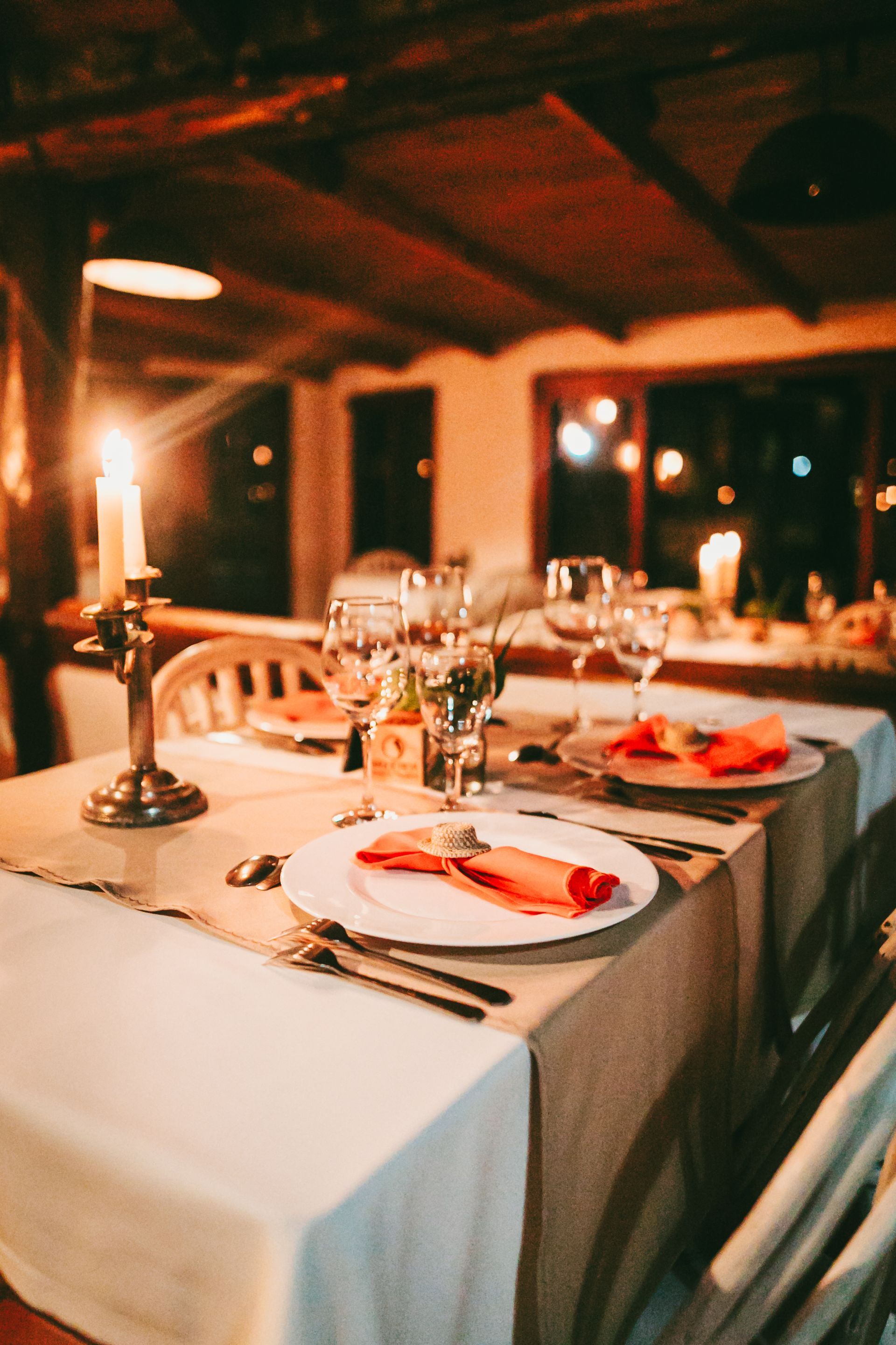Mesa de comedor iluminada con velas, puesta con manteles blancos, copas de vino y servilletas rojas en un restaurante acogedor.