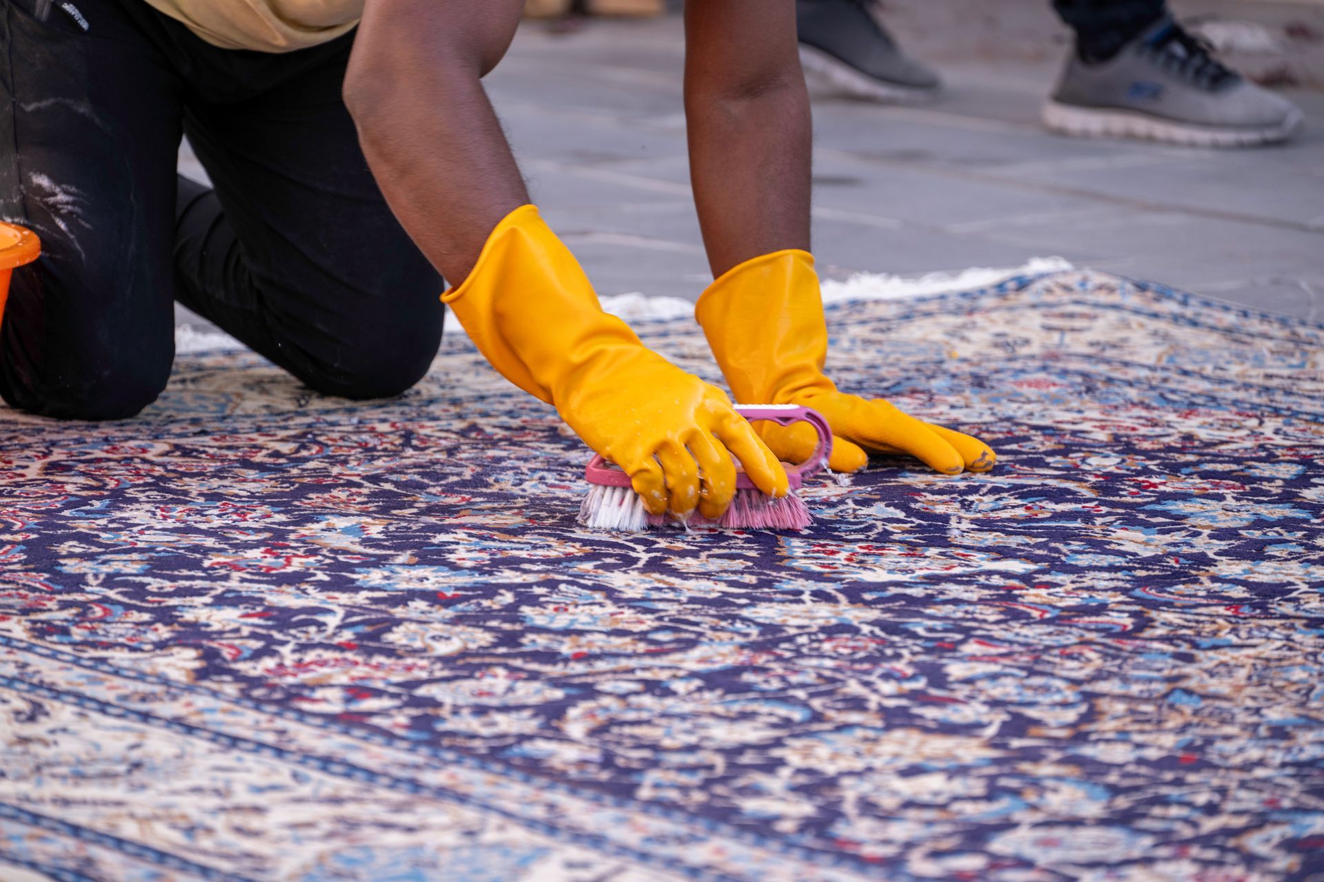 Person in yellow gloves scrubs a patterned rug with a brush and bucket.
