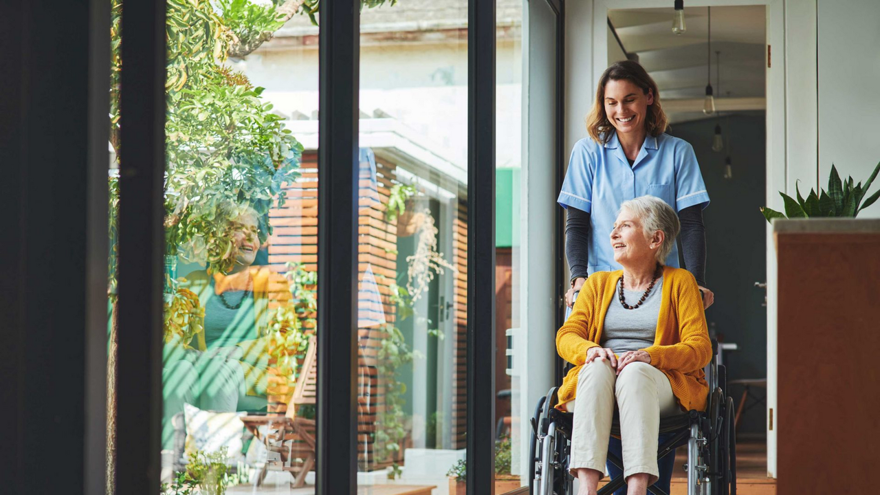 Woman with cane assisted by caregiver on a porch; caregiver's hand on her back for support.