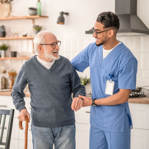 Caregiver in blue scrubs assists older person outdoors, smiling.