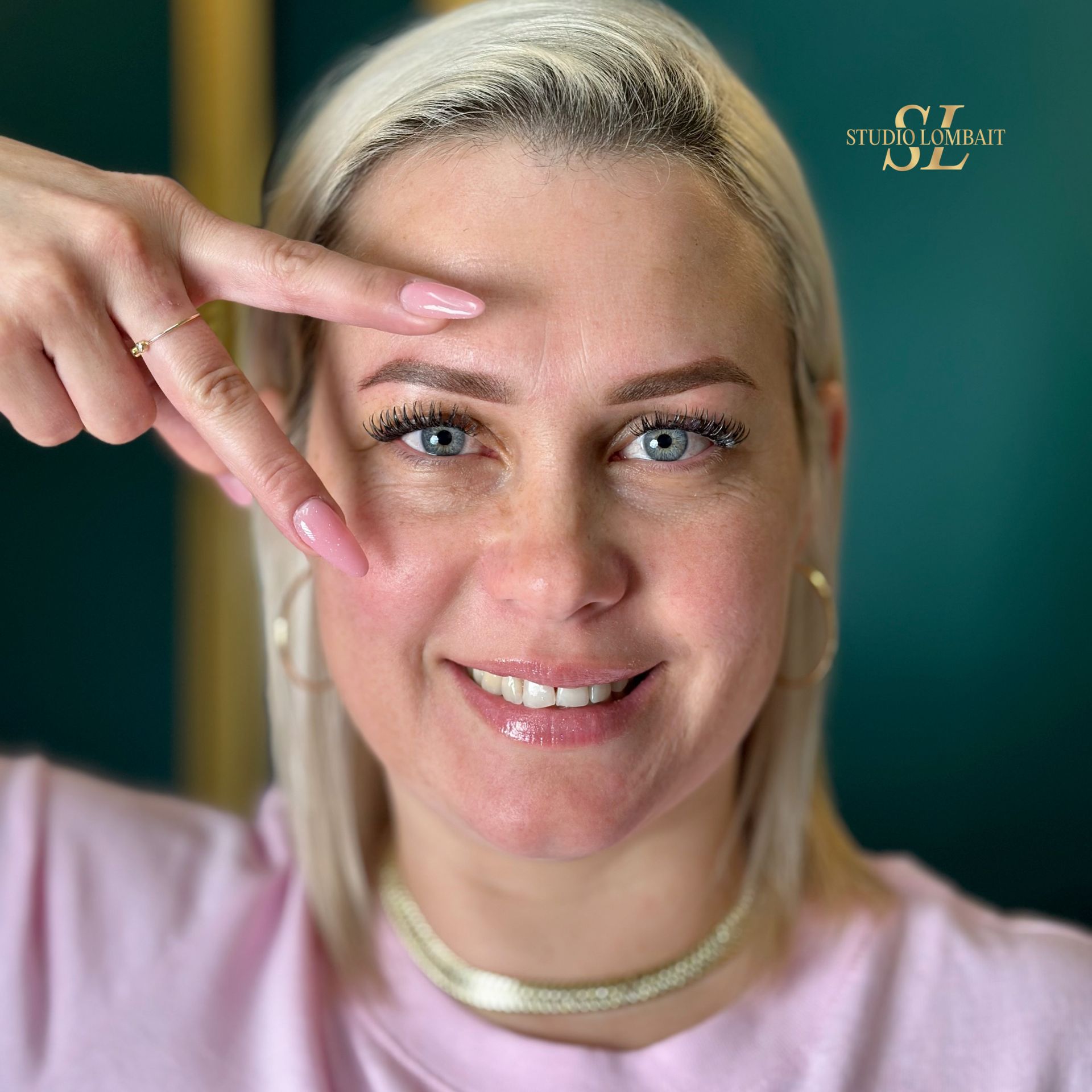 A woman with blonde hair is smiling and pointing at her eyebrows