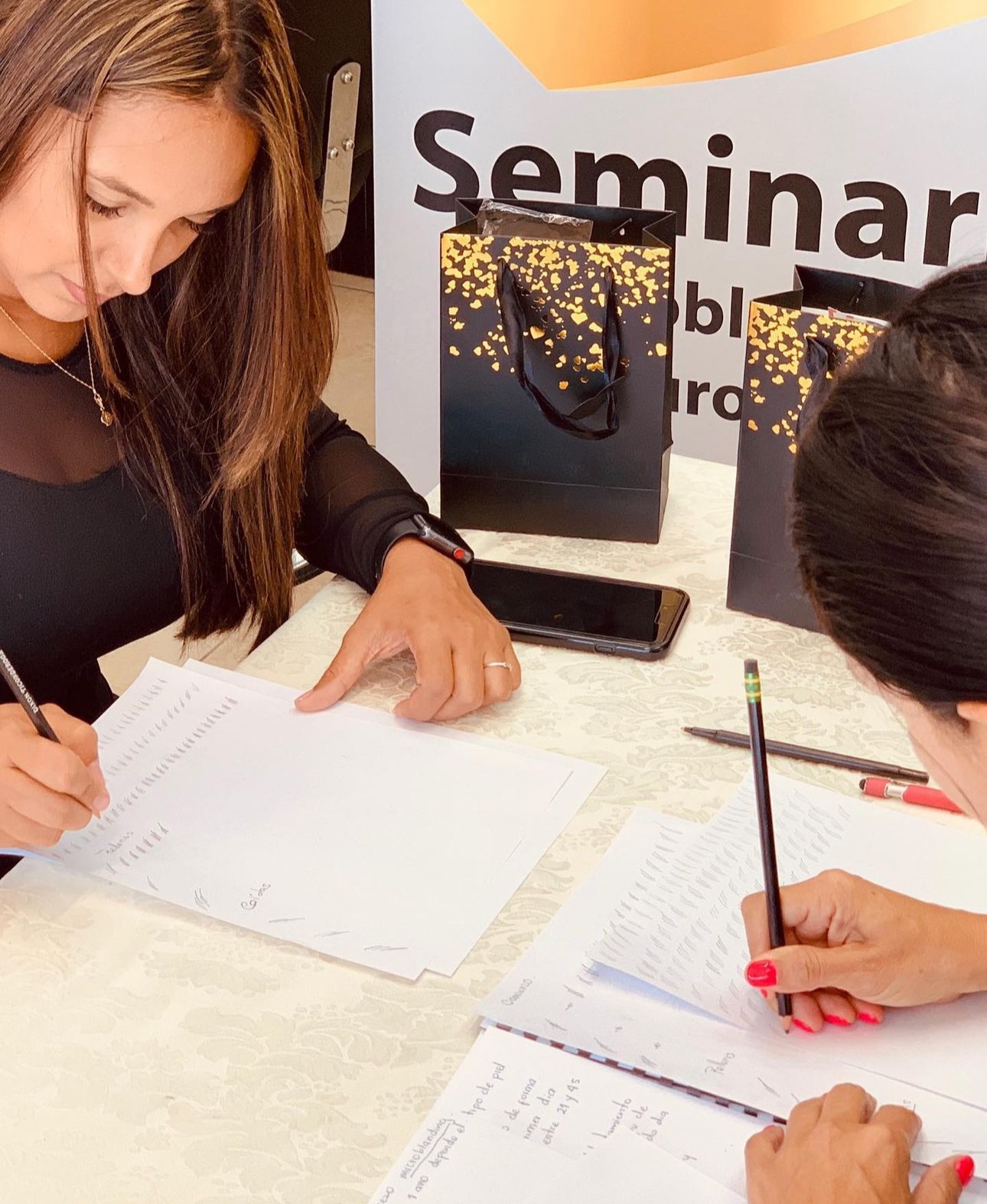 Two women are sitting at a table writing in notebooks.