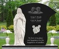 A black granite memorial headstone featuring a white marble statue of a praying figure, floral vases, and inscriptions.