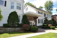 A white, two-story building with a projecting covered entrance, dark shutters, manicured shrubs, and a grassy lawn.