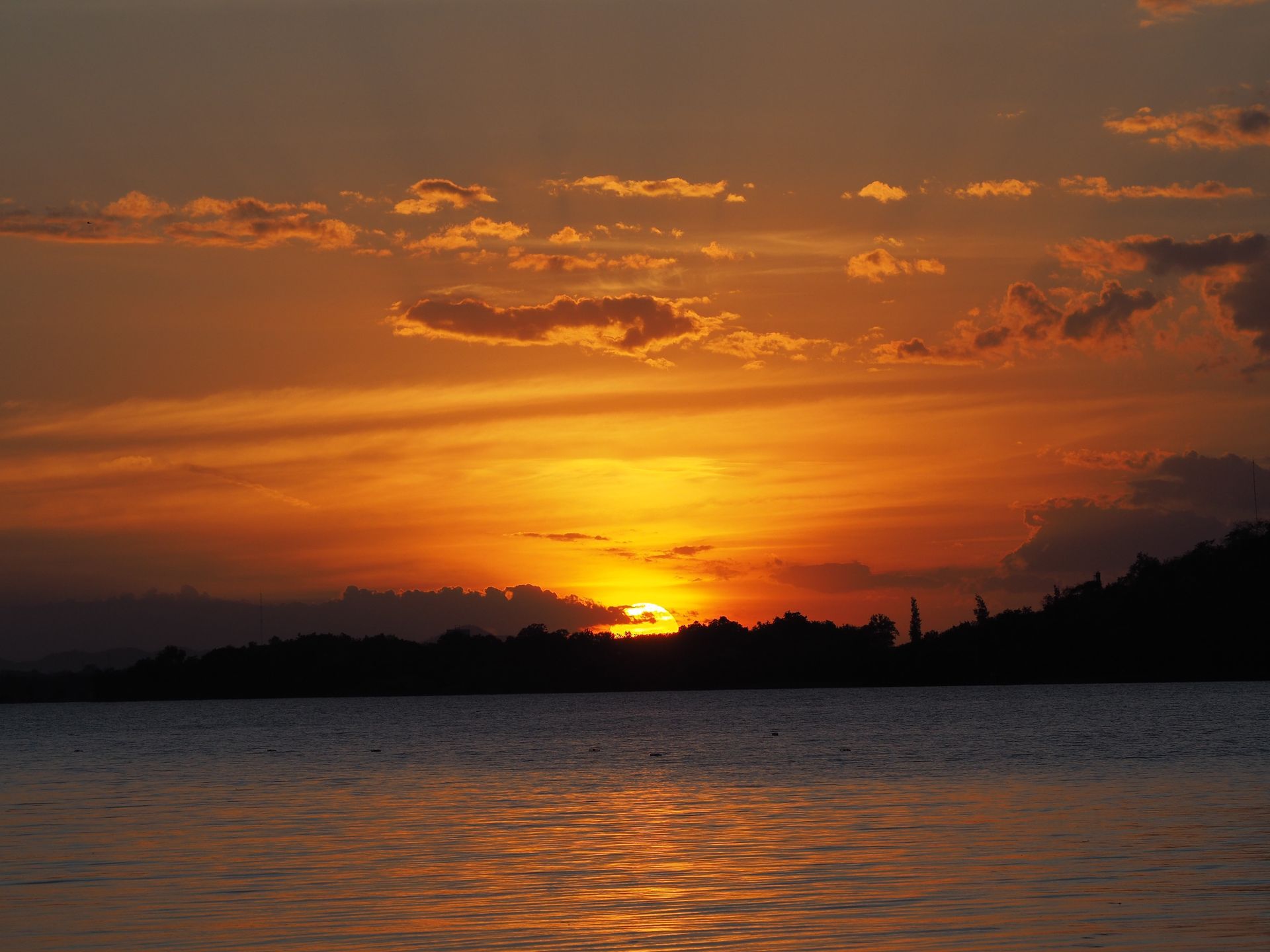 The sun sets behind a dark silhouette of trees and hills over a calm body of water with vibrant orange and gold clouds.