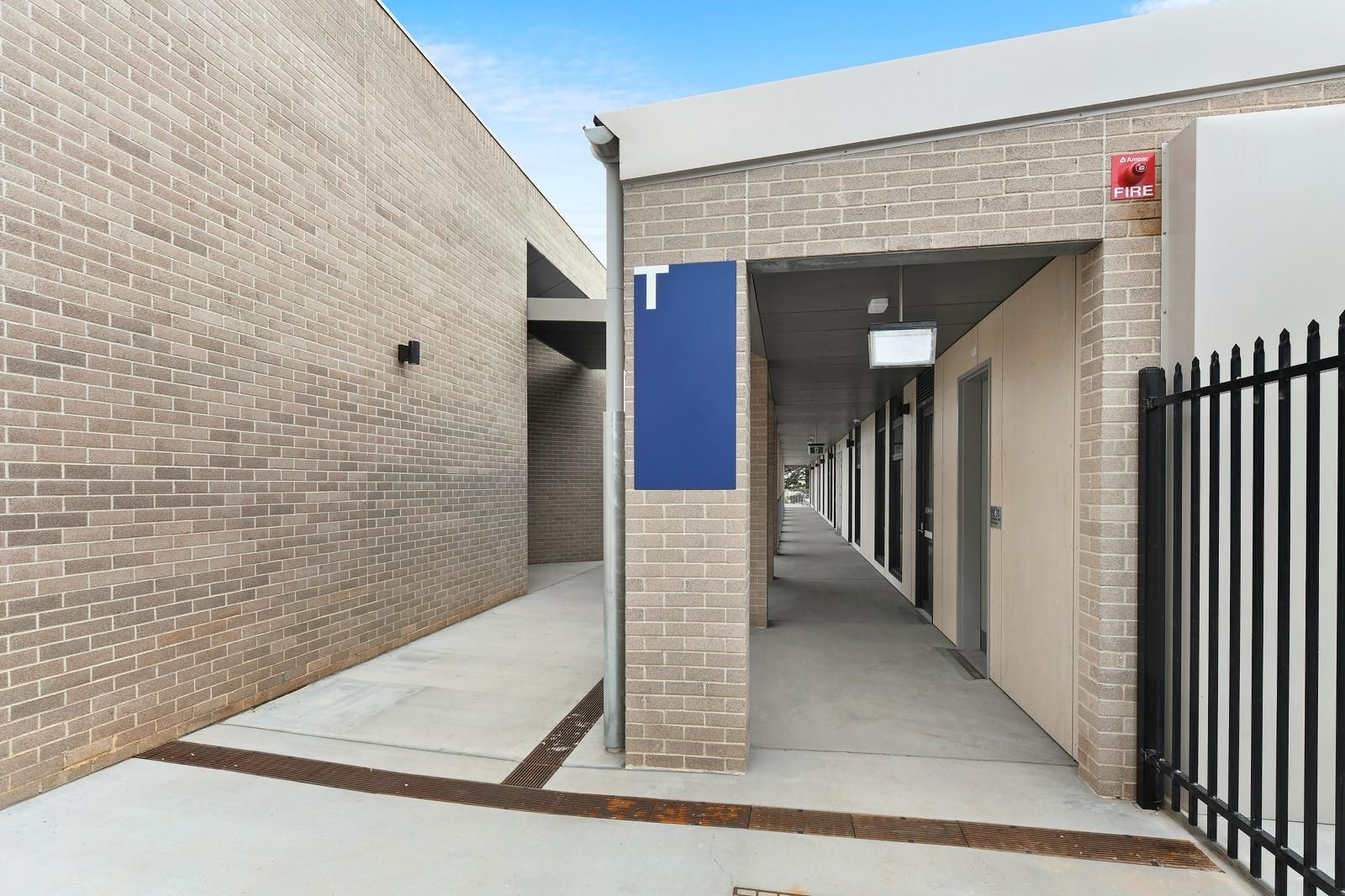 Covered outdoor corridor beside tan brick buildings, with a blue sign and black gate. — Millers Masonry In Port Macquarie, NSW