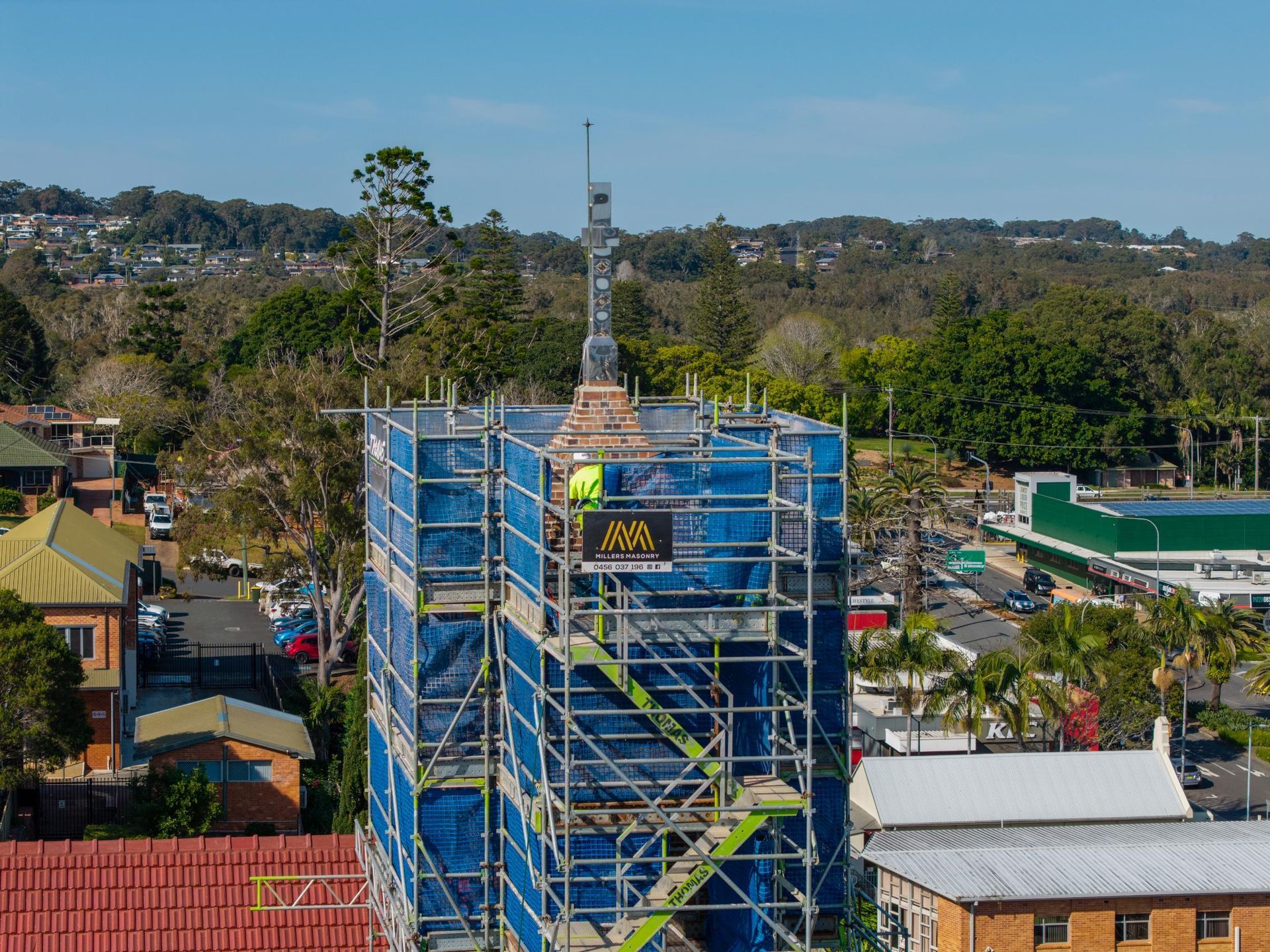 Scaffolding Surrounds a Building's Spire with Workers on A Platform; Blue Tarp, Green Trees — Millers Masonry In Port Macquarie, NSW