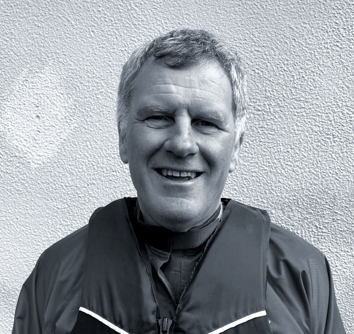 Man in a life jacket, smiling, black collar, outdoor setting, grayscale.
