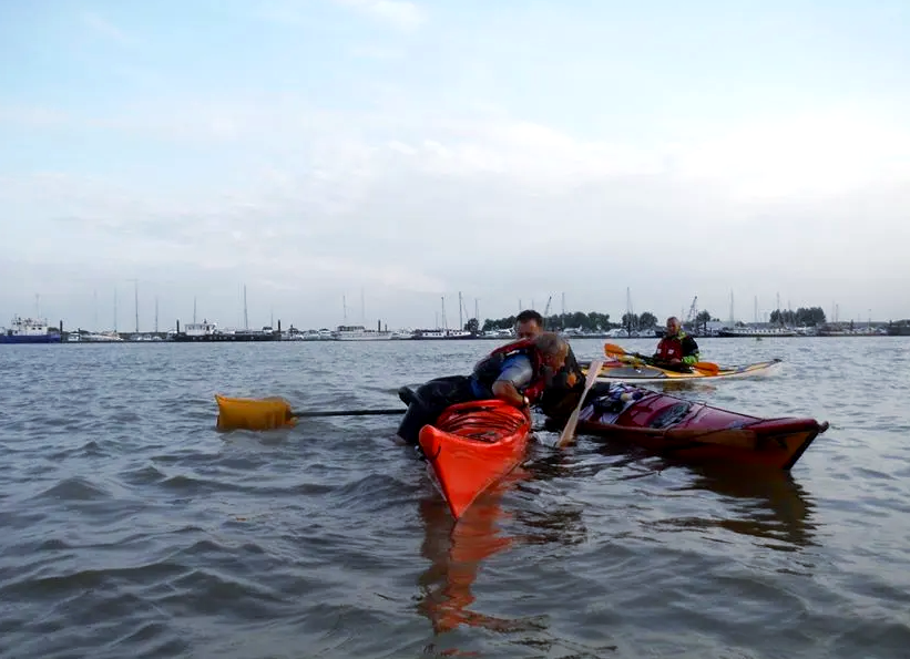 Kayakers near a harbor with boats. One red kayak, two people helping load gear, orange buoy. Cloudy sky.