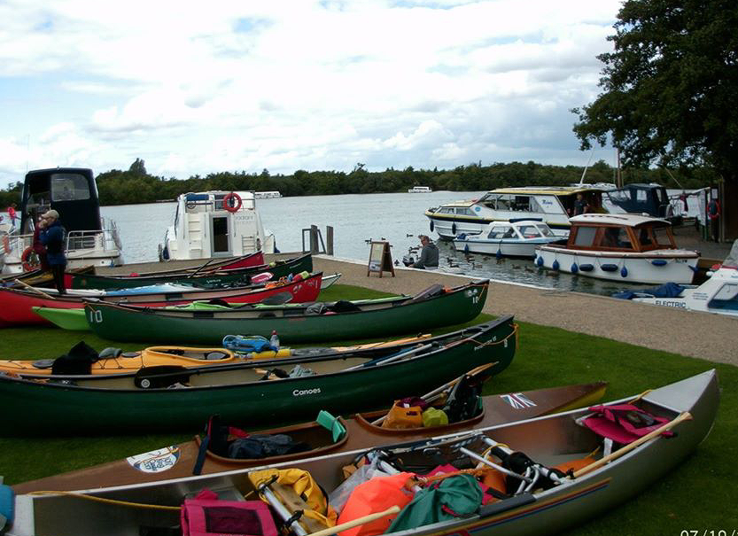 Canoes and boats lined up on a grassy shore next to a body of water under a cloudy sky.