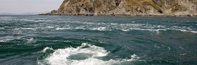 Whirlpool in ocean near a rocky, brown landmass under an overcast sky.