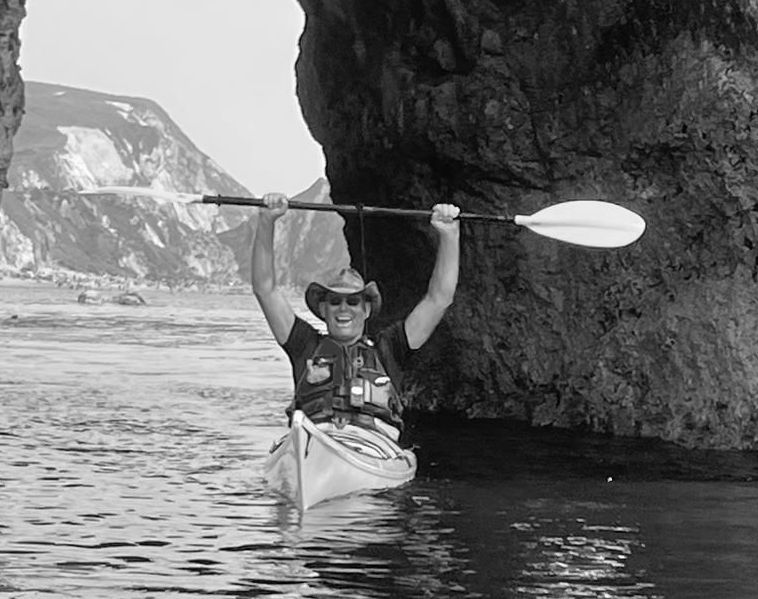 Man in kayak raises paddle, smiles under rock arch.