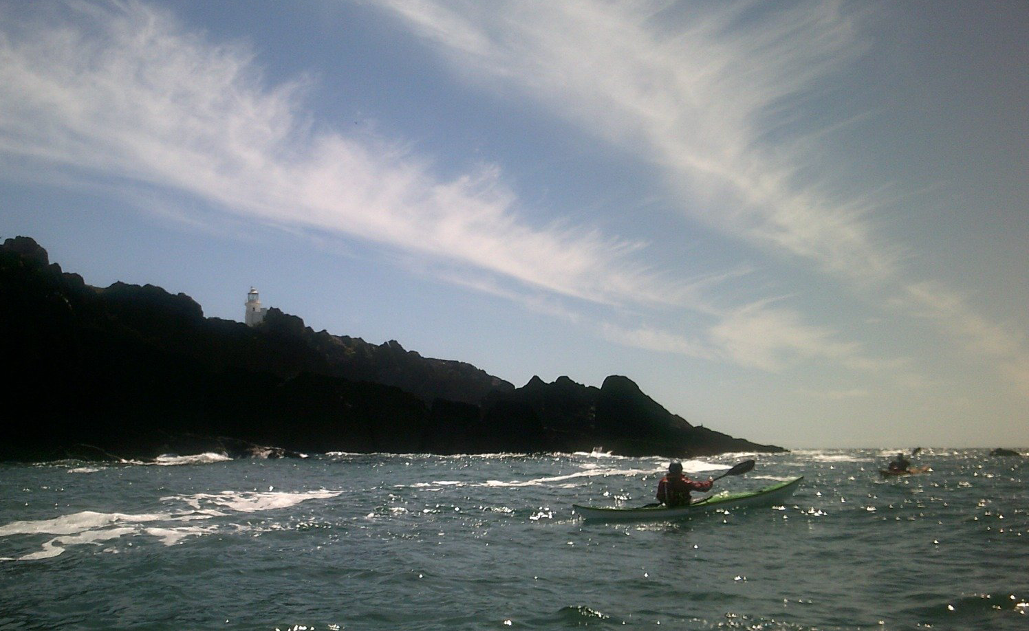 Kayakers on ocean near dark cliff, under a partly cloudy sky with a white lighthouse on top.