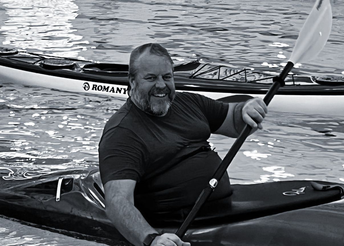 Man in kayak, smiling, holding paddle, water in background. Another kayak is behind him.