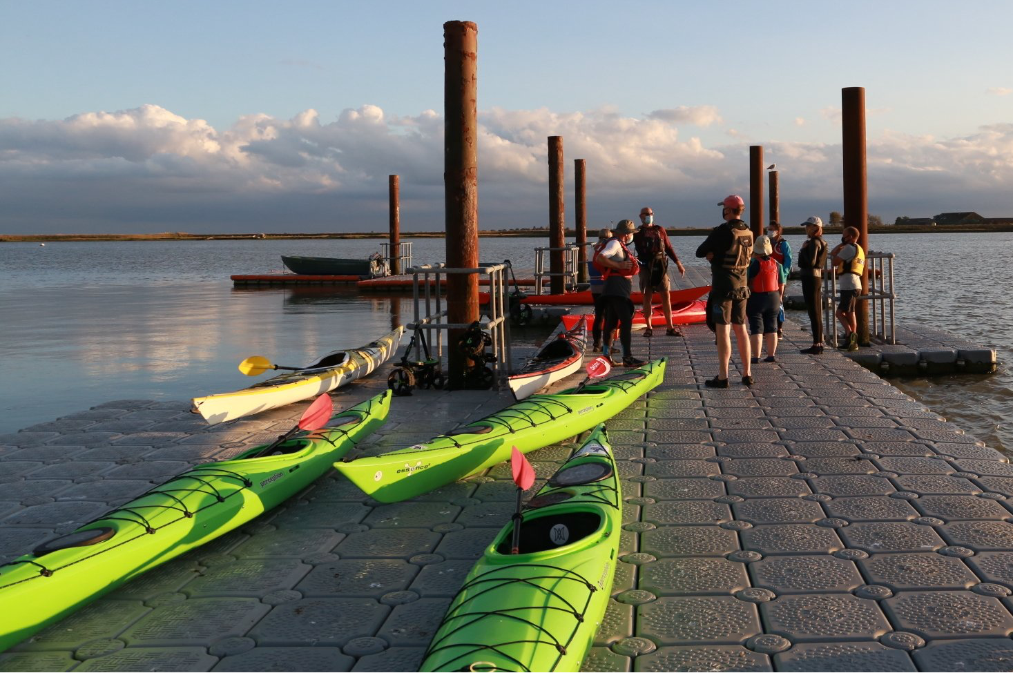 Kayaks lined up on a dock, people standing nearby. Setting is a lake at sunset.