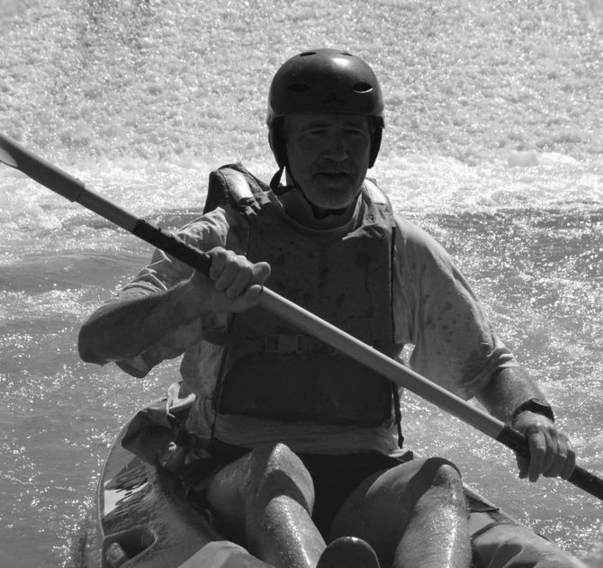 Man kayaking in whitewater, wearing a helmet and life vest, holding a paddle.