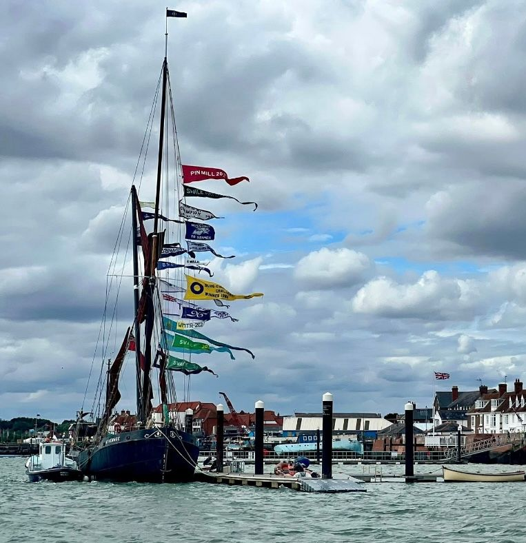Sailboat docked with many colorful flags, against a cloudy sky, buildings in the background.