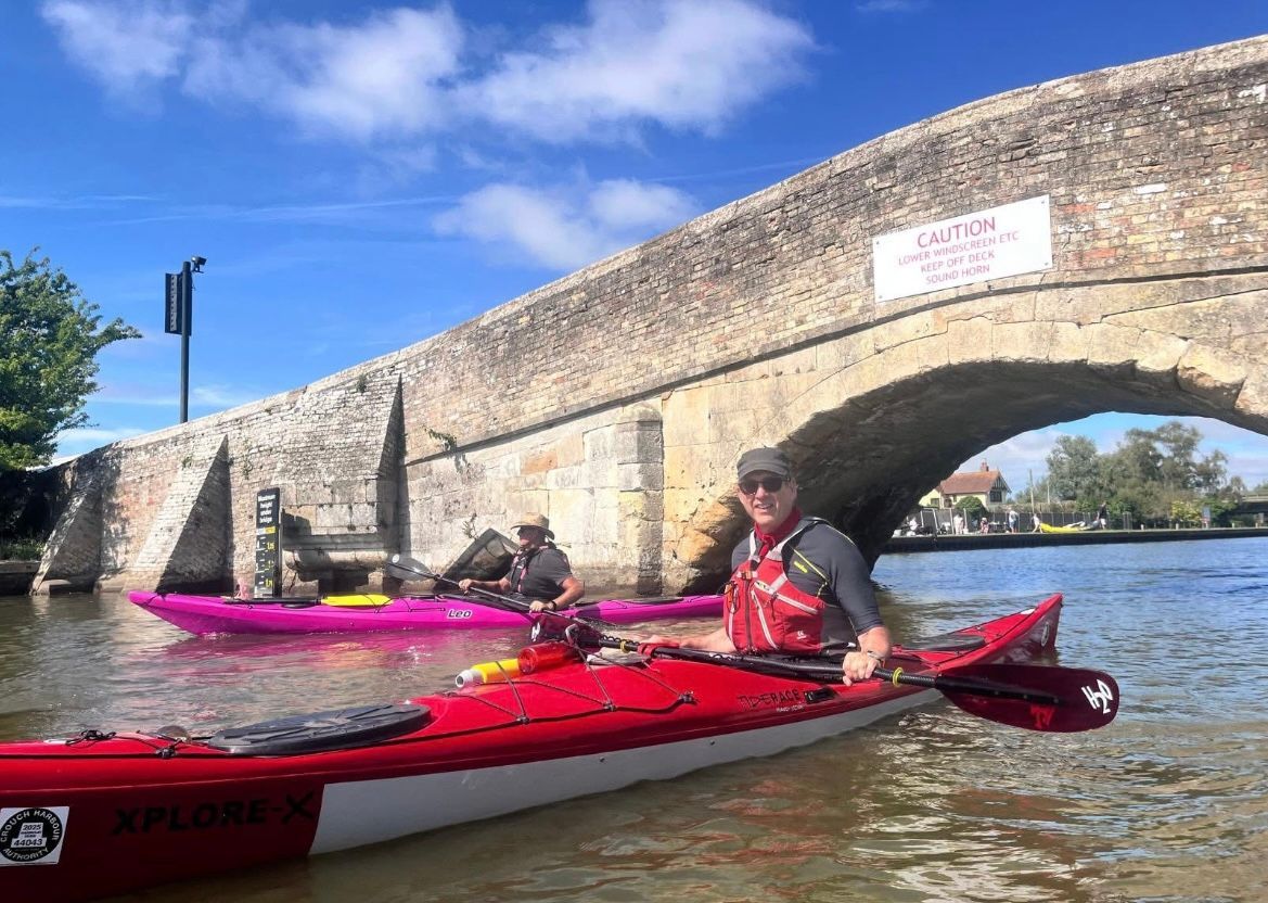 Two people kayaking under an old stone bridge on a sunny day. Red and pink kayaks.