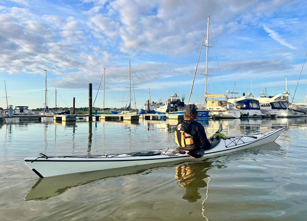 Person kayaking in calm water, harbor with boats in background, blue sky.