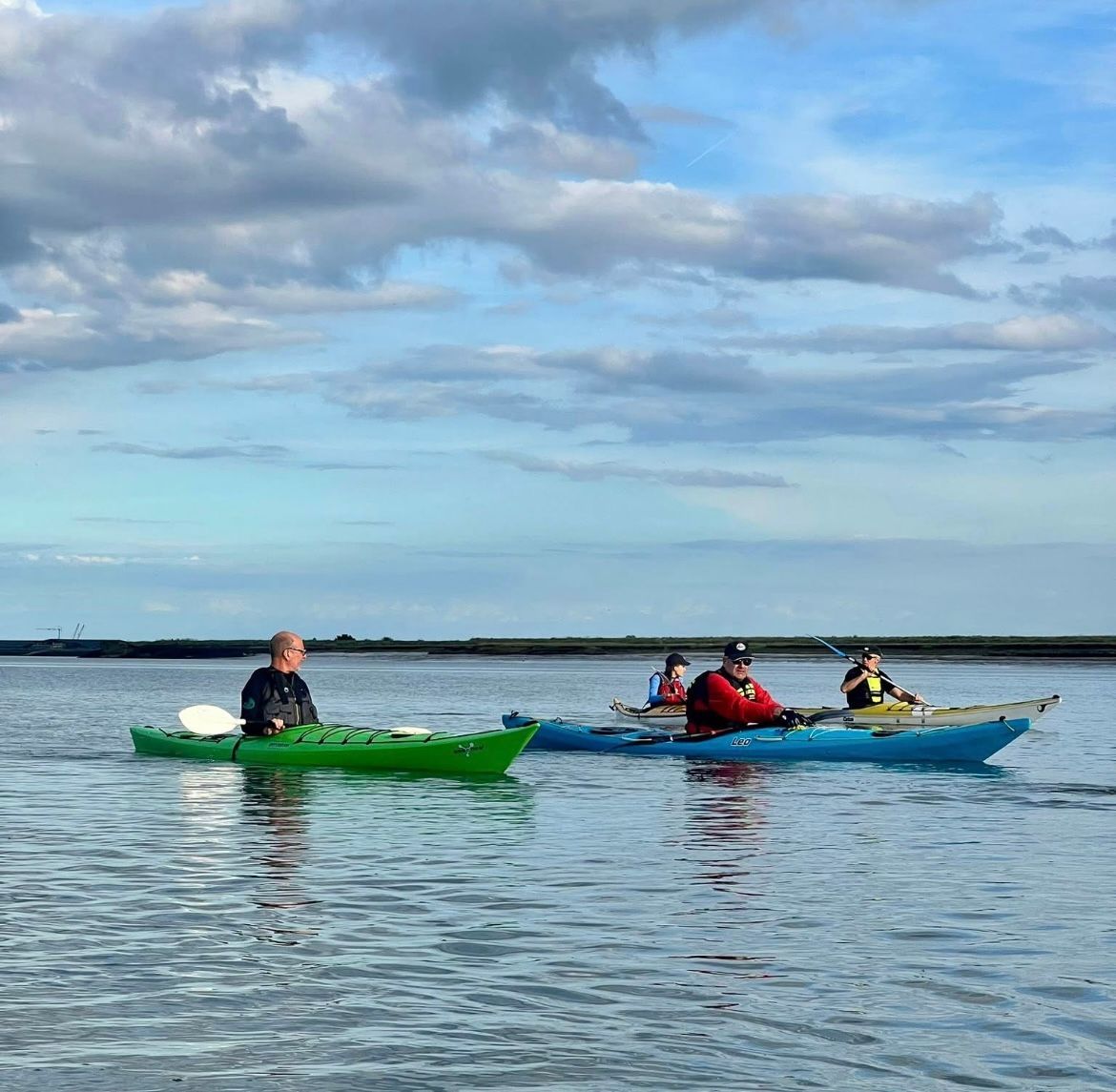 Kayakers on calm water under a cloudy sky. Green, blue, and yellow kayaks with paddlers near a coastline.