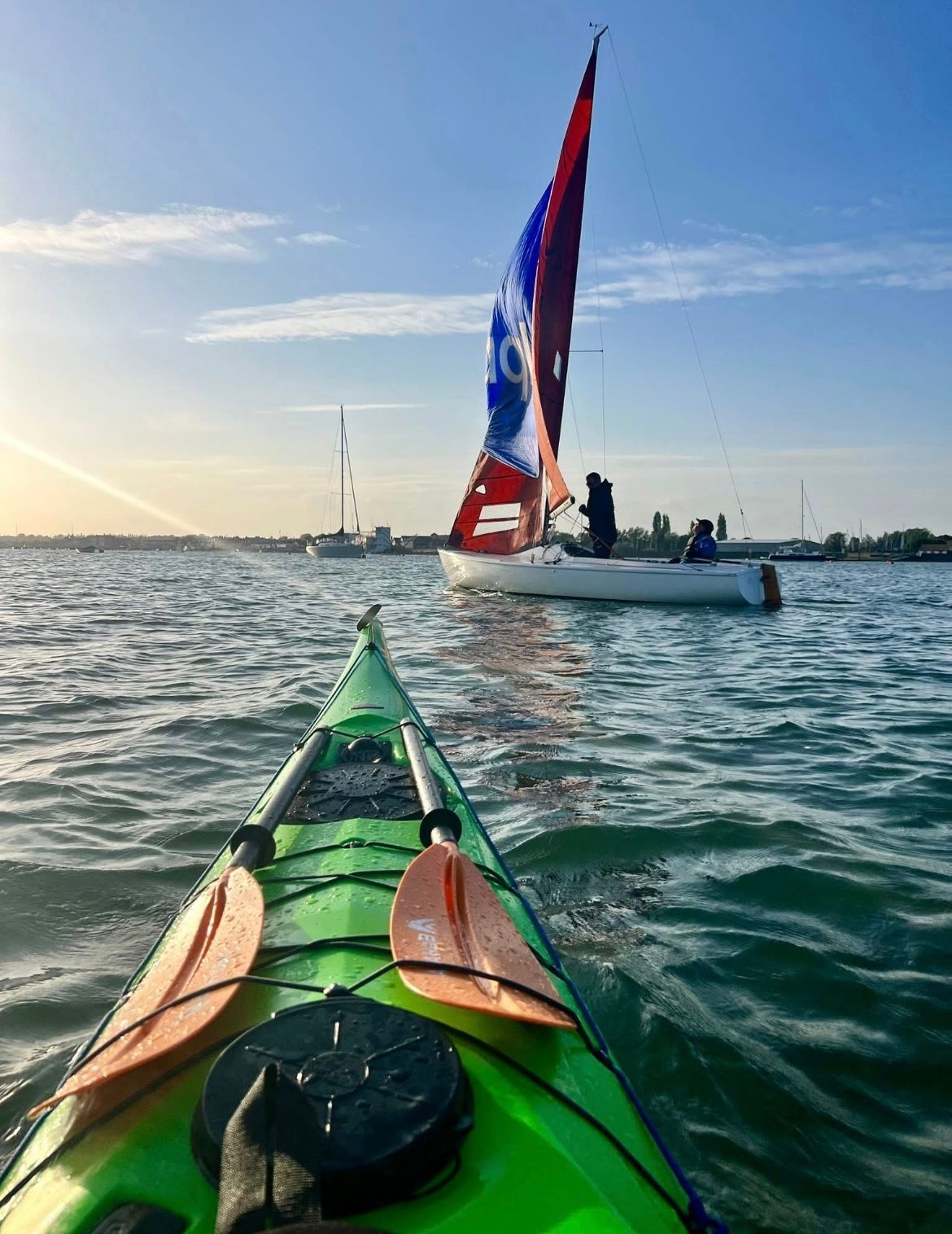 Green kayak in the foreground, sailing boat in background with red, blue, and white sail. Water and sky.
