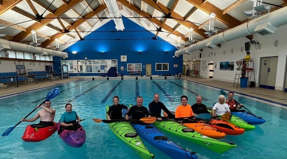 People in kayaks in a turquoise pool. Inside a building with blue walls, ceiling beams.