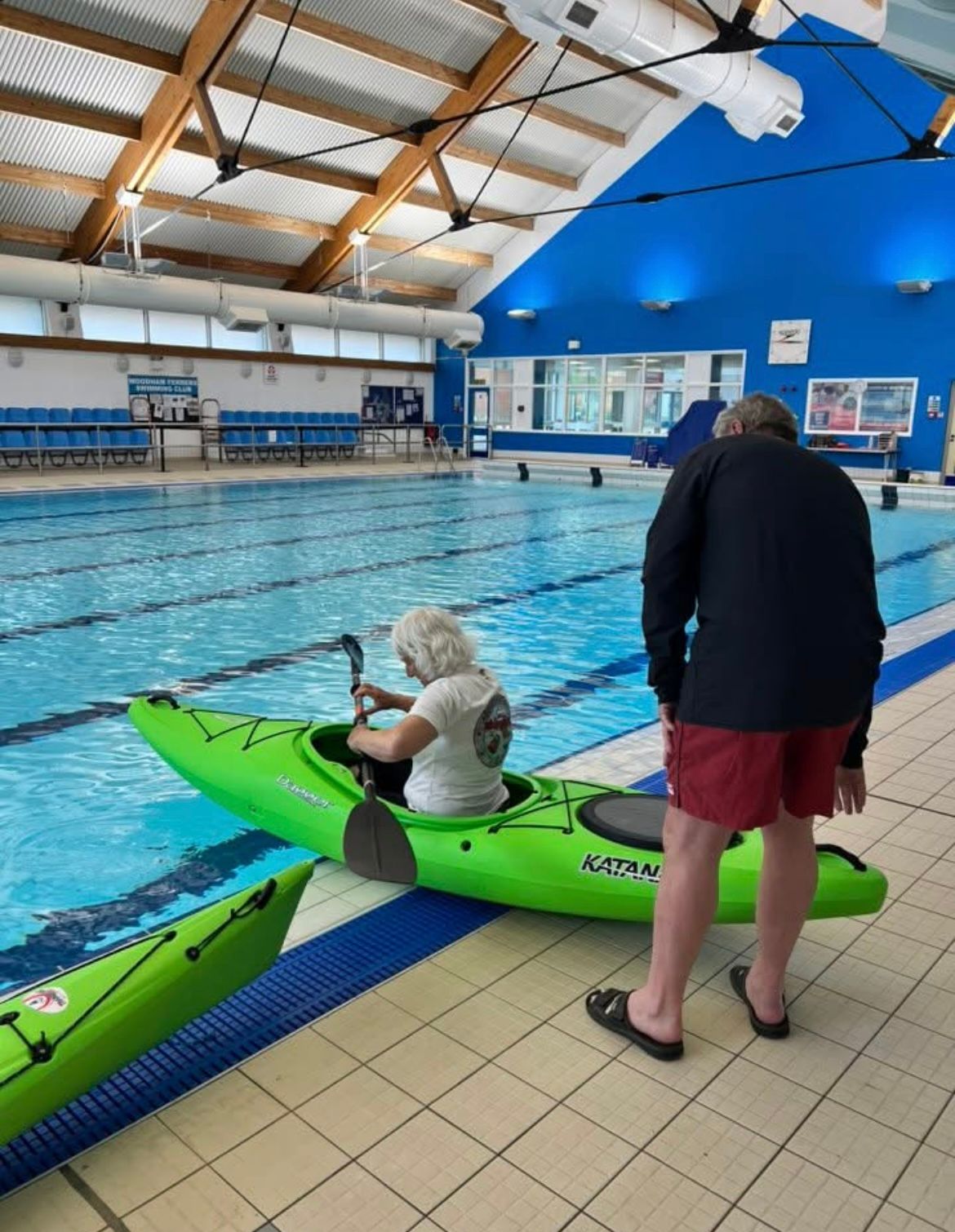 A person in a kayak in an indoor pool, being assisted by another. The kayak is green.