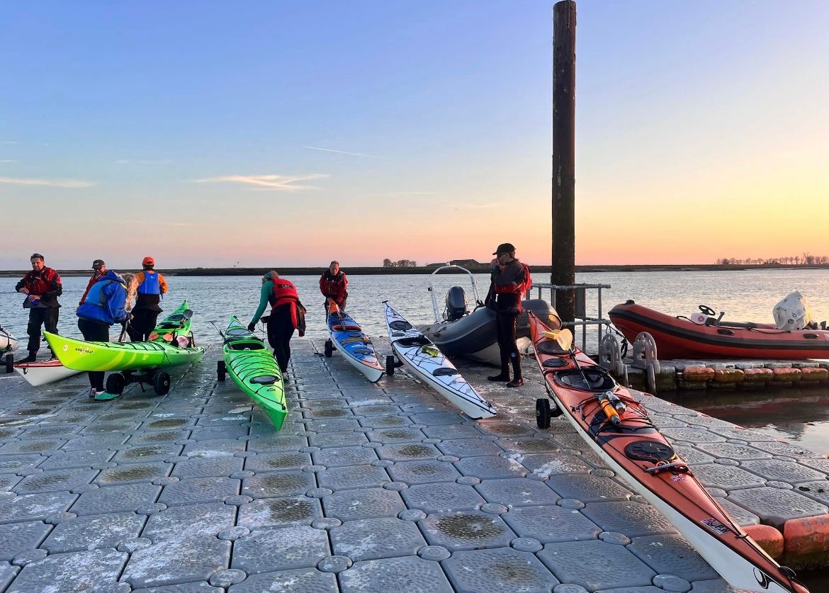 Kayakers preparing to launch from a dock at sunrise, various kayaks and an inflatable boat present.