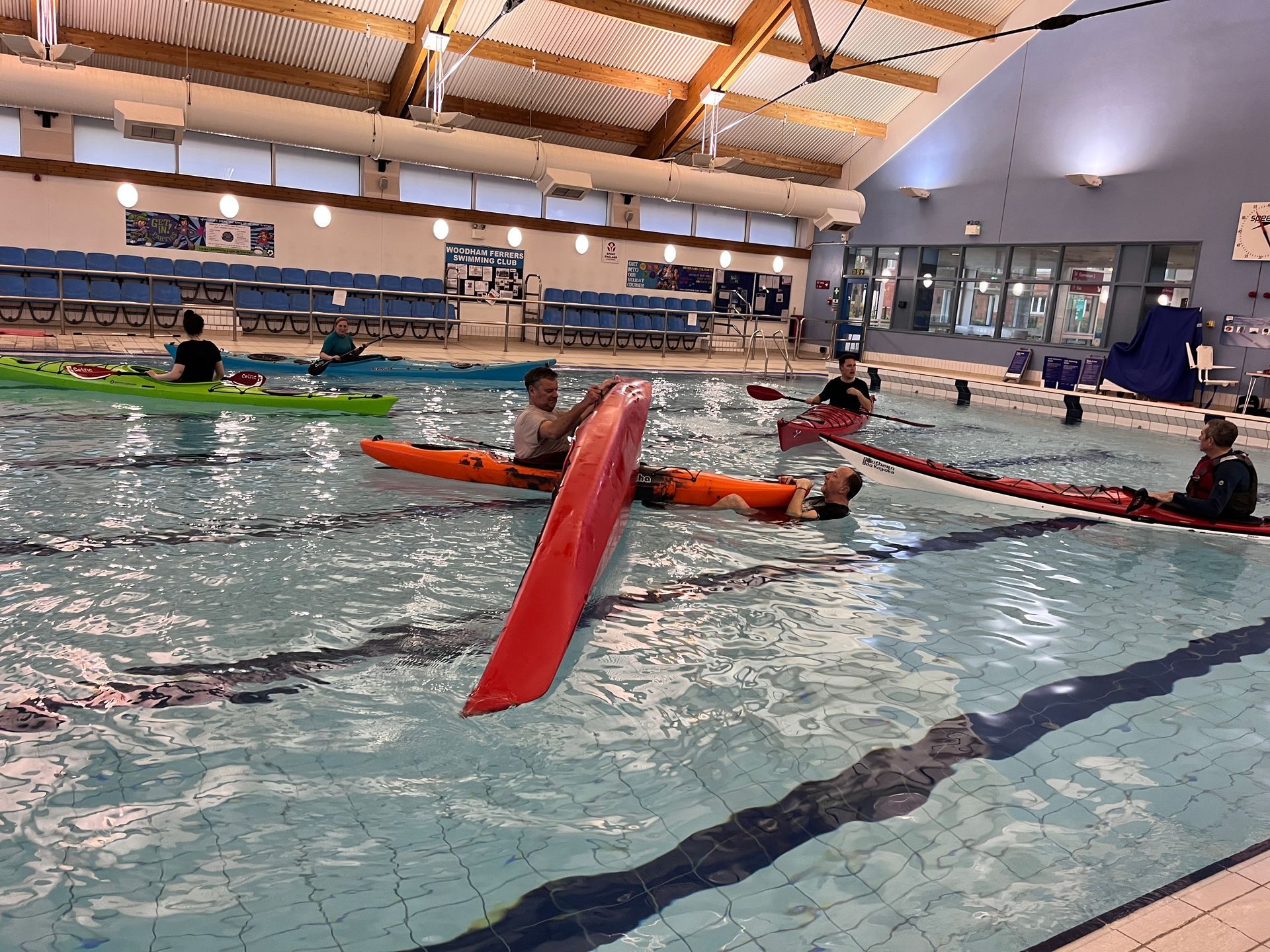 Kayakers in a swimming pool; red kayak is upright, others are paddling. Indoor pool setting.