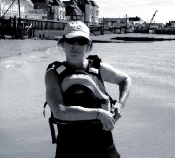 Woman in a life vest and sunglasses stands near water, buildings in the background.