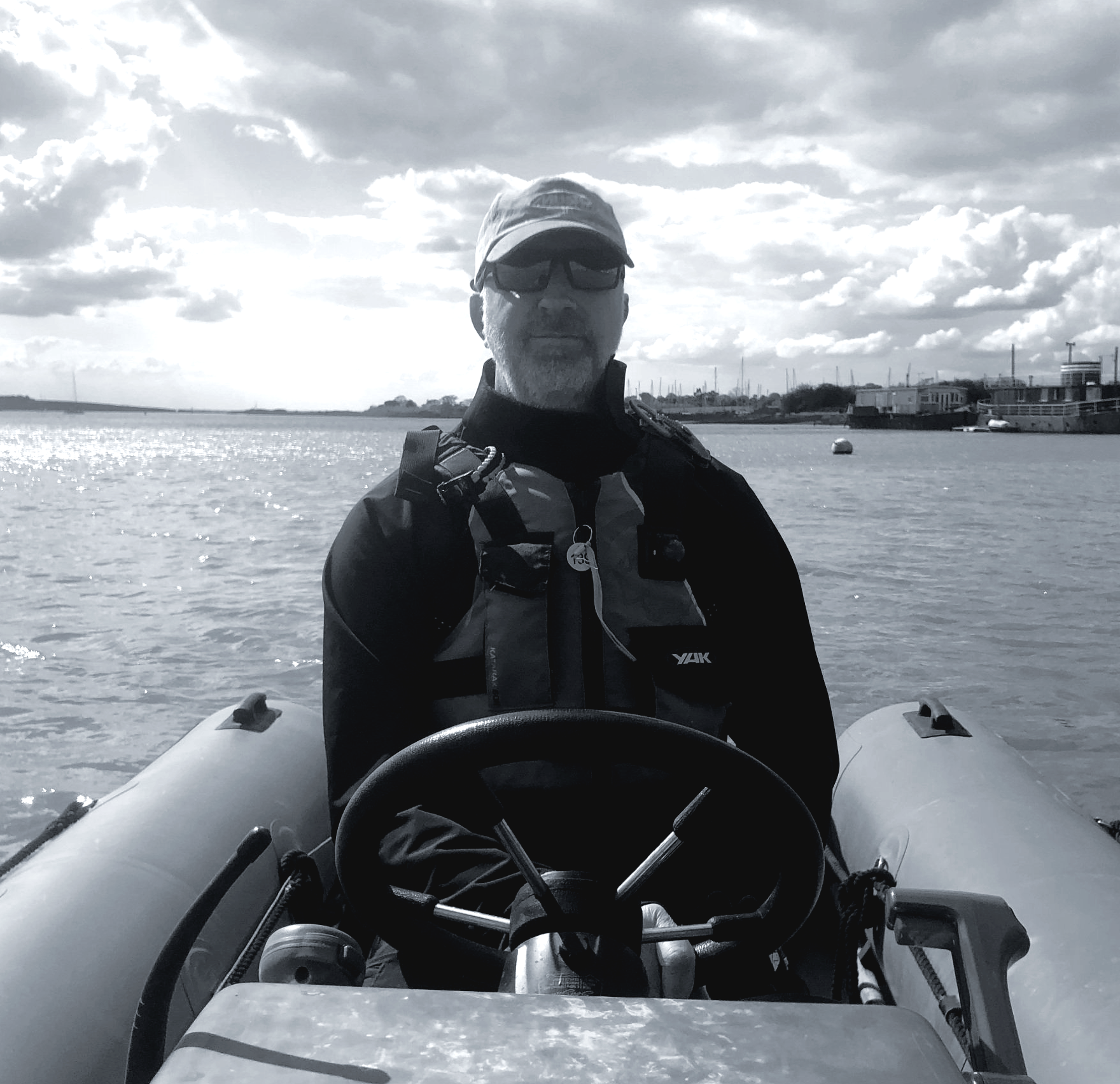 Man in a boat, wearing a cap and life jacket, holding a steering wheel, on the water.
