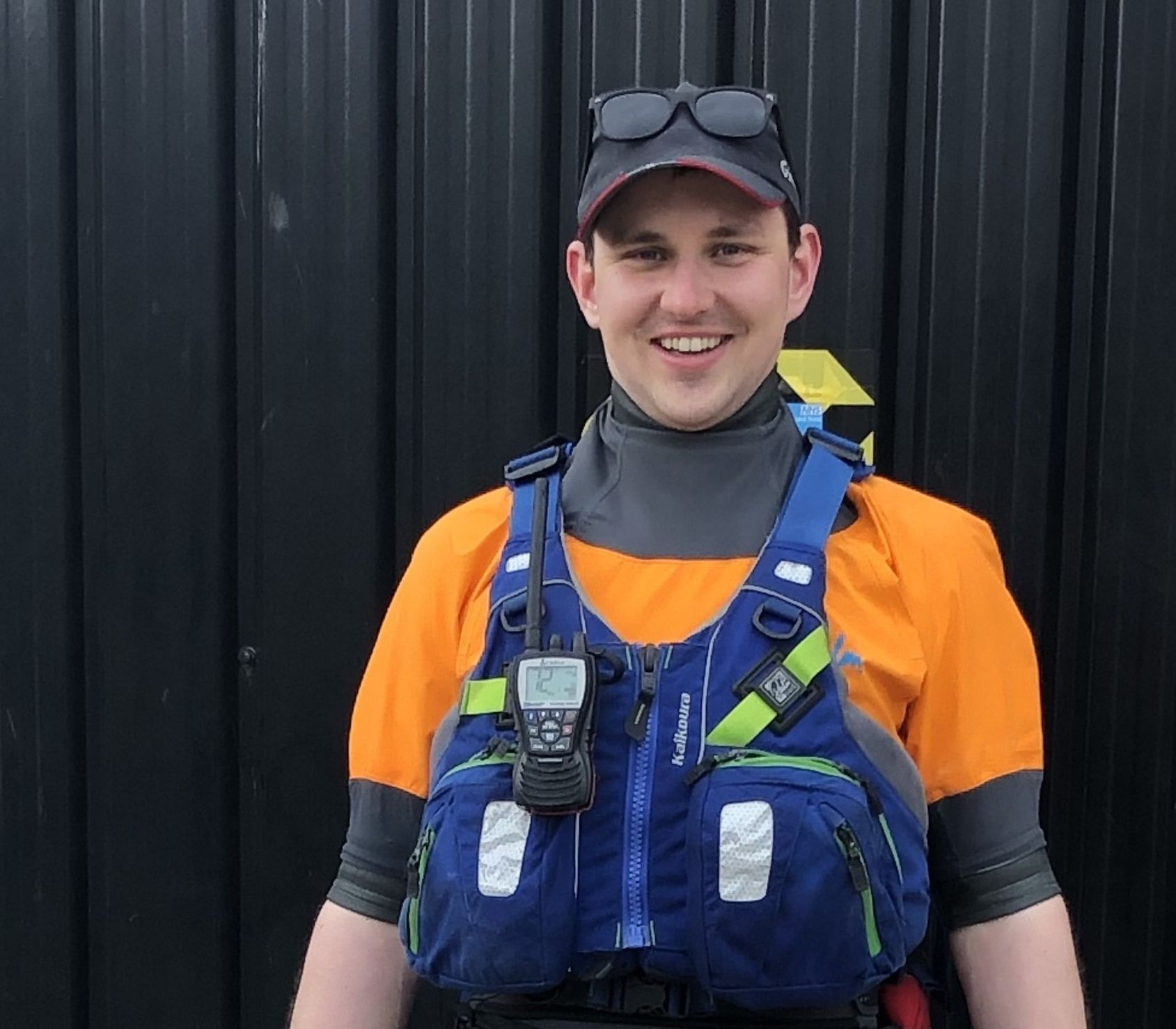 Man wearing a blue life vest, orange shirt, and black hat smiles, standing in front of a black wall.