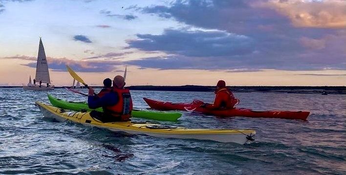 Kayakers paddling on water with sailboat in the distance, against a cloudy sunset sky.