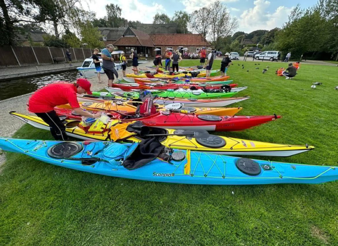 Kayaks lined up on grass, people preparing. Red shirt, various boat colors. Outdoors, sunny day.