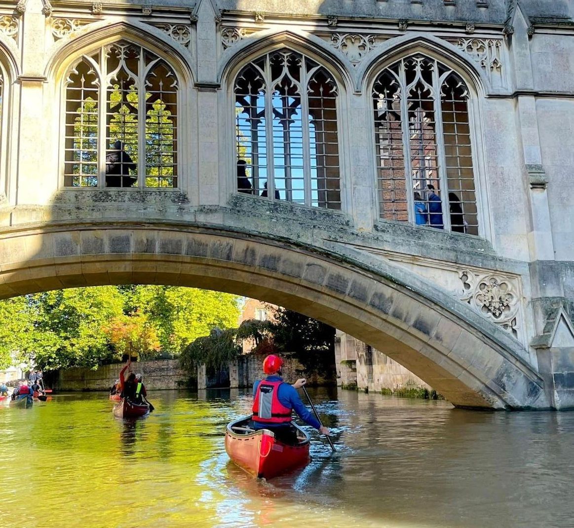 Canoeists on a river under a stone bridge with arched windows. Sunlight reflects on the water.